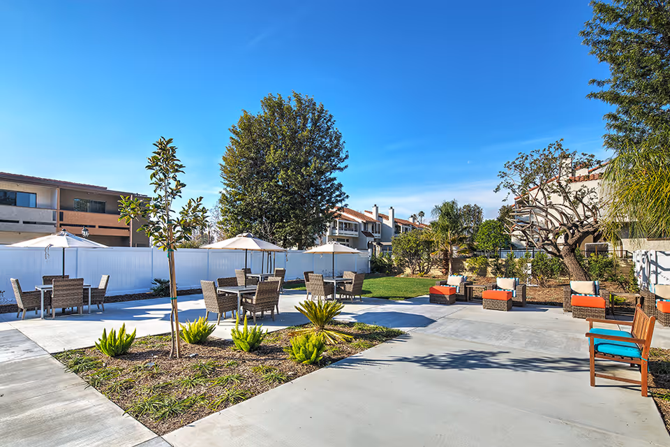 Outdoor patio area at a senior living facility with multiple tables and chairs under umbrellas, surrounded by greenery and residential buildings under a clear blue sky.