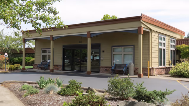 Front entrance of a single-story nursing and rehabilitation center with a covered drop-off area, glass double doors, benches, and landscaped plantings.