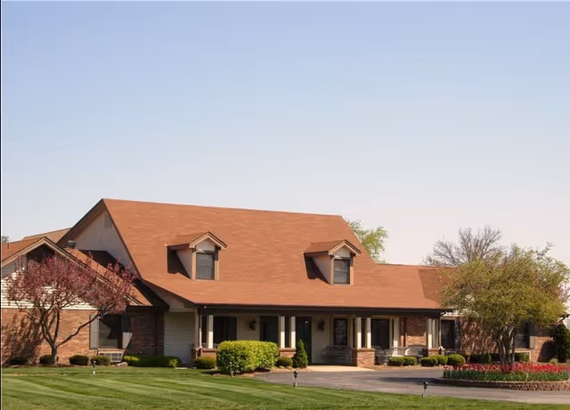 A single-story building with a brown roof and brick and beige siding exterior, surrounded by neatly trimmed bushes, trees, and a well-maintained lawn under a clear blue sky.