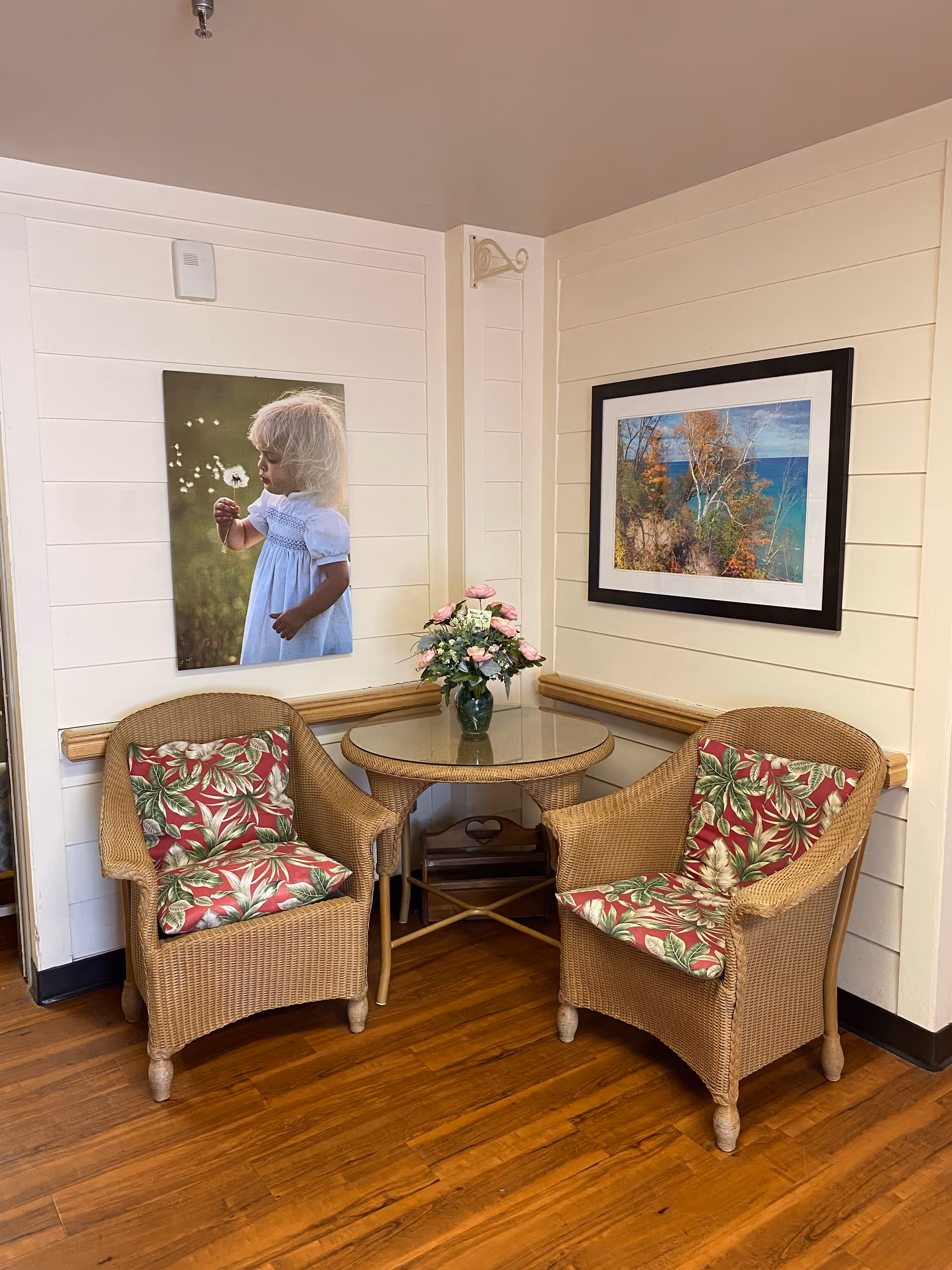 A cozy corner with two wicker chairs featuring floral cushions, a round glass-top wicker table with a vase of pink and white flowers, and two framed pictures on the cream-colored paneled walls. The floor is wooden.