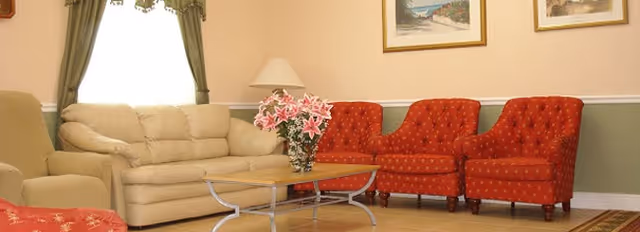Cozy living room with a beige sofa, three red upholstered chairs, a glass-top coffee table holding a vase of flowers, lamp, and framed paintings.