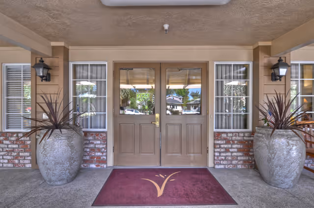 Double wooden doors frame a brick entrance with tall planters and a maroon welcome mat.