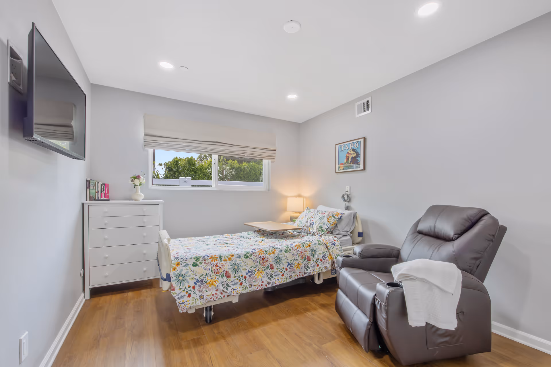 A bright, modern bedroom in a senior living facility with a floral bedspread and recliner.