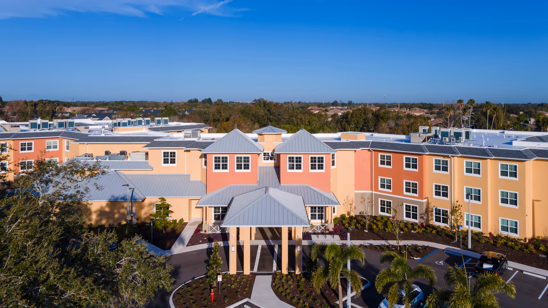 Aerial view of a colorful three-story senior living building with a covered porte-cochère entrance, palm trees, and a parking area.
