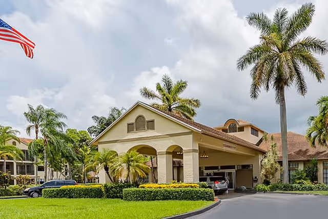 Exterior view of a senior living facility building with a covered entrance, surrounded by palm trees and well-maintained landscaping under a cloudy sky. An American flag is visible on the left side, and a car is parked near the entrance.