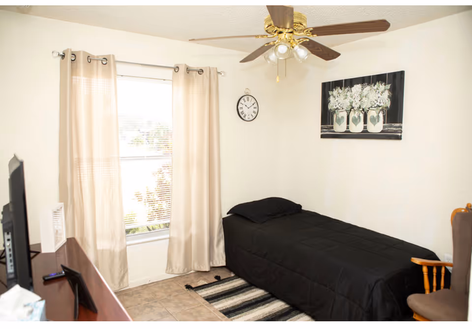 A small bedroom with a black bed, ceiling fan, window with beige curtains, and floral art.