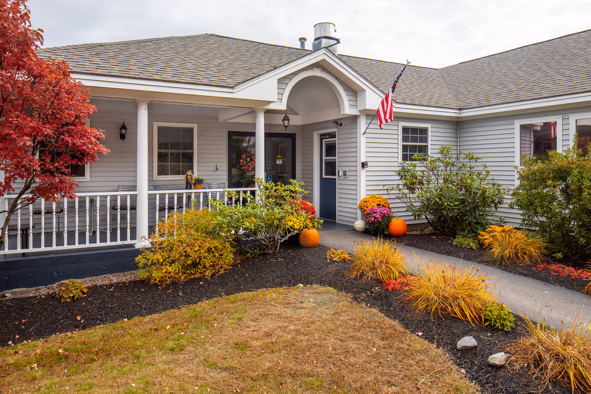 Front entrance of a single-story senior living building with a covered porch, American flag, fall plants and pumpkins.