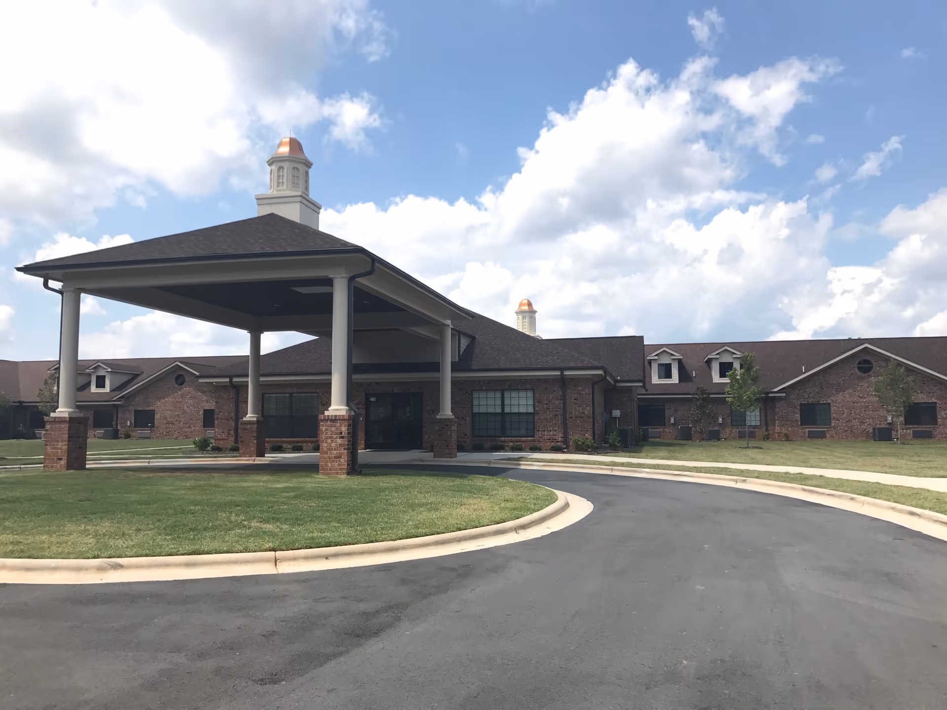 Front exterior view of a single-story brick building with a covered entrance supported by columns, a circular driveway, and a well-maintained lawn under a partly cloudy sky.
