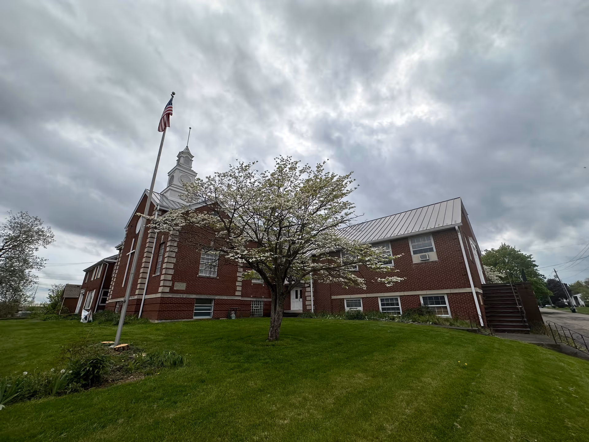 Red brick building with white trim, blooming tree, flagpole with American flag, under cloudy sky.