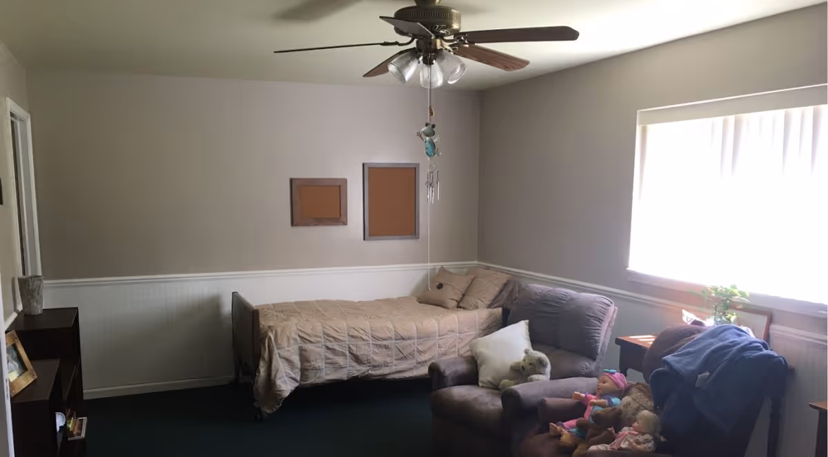 A modest bedroom with a single bed against the wall, a ceiling fan with lights, a cushioned armchair with pillows and stuffed animals, a window with blinds letting in natural light, two empty cork boards on the wall, and a small bookshelf with a framed photo and other items.
