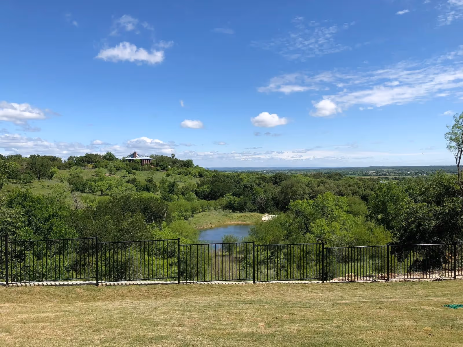 Grassy yard with a black metal fence overlooking a wooded valley with a small pond, a distant building on a hill, and a blue sky with scattered clouds.