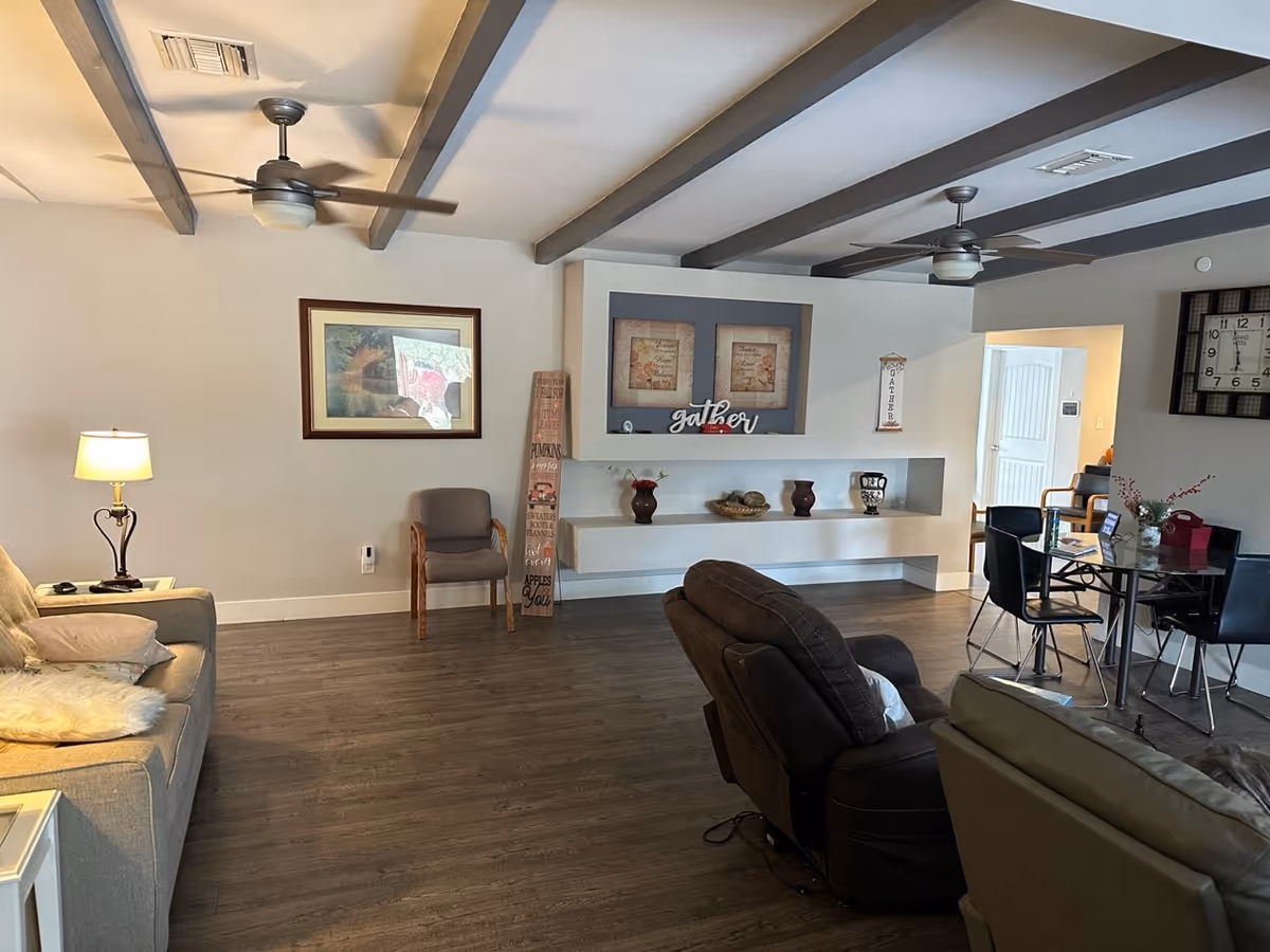 A cozy living room area with wooden flooring, a beige sofa with cushions and a lamp on a side table, a brown recliner chair, and a gray chair against the wall. The wall features a framed picture, decorative shelves with vases and a basket, and the word 'gather' displayed. There is a glass dining table with black chairs and a large wall clock. Ceiling fans with lights hang from the ceiling with exposed beams.