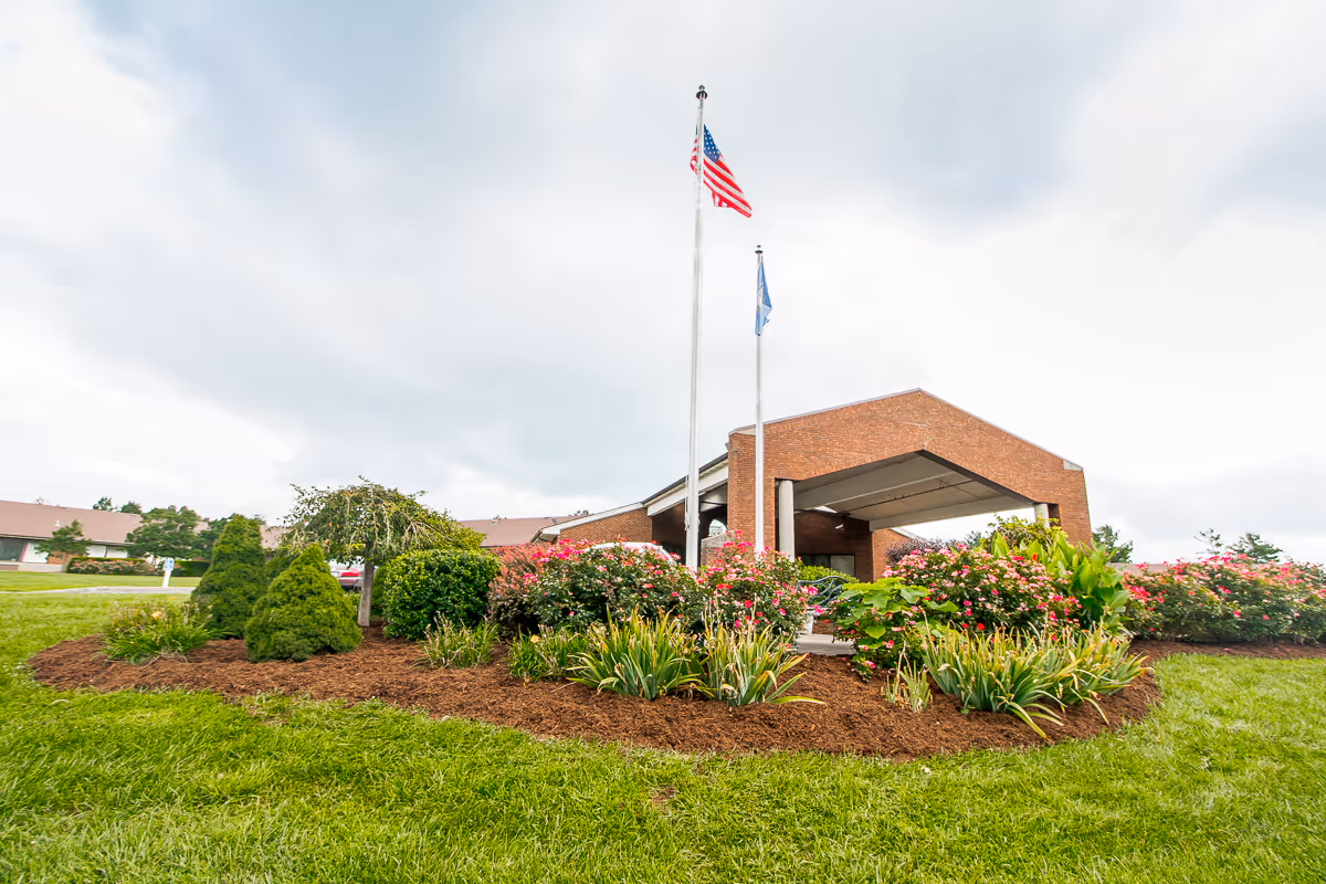 Brick senior living facility exterior with landscaped garden, flagpoles, and covered entrance.
