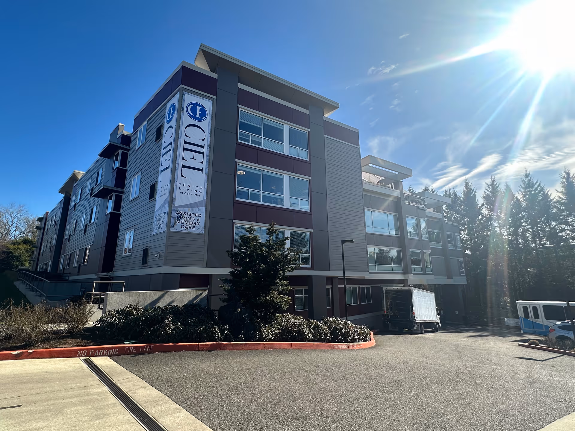 Exterior view of a modern multi-story senior living facility building named Ciel of Cedar Mill under a clear blue sky with sunlight shining brightly. There are trees in the background and a parking area with vehicles in front.