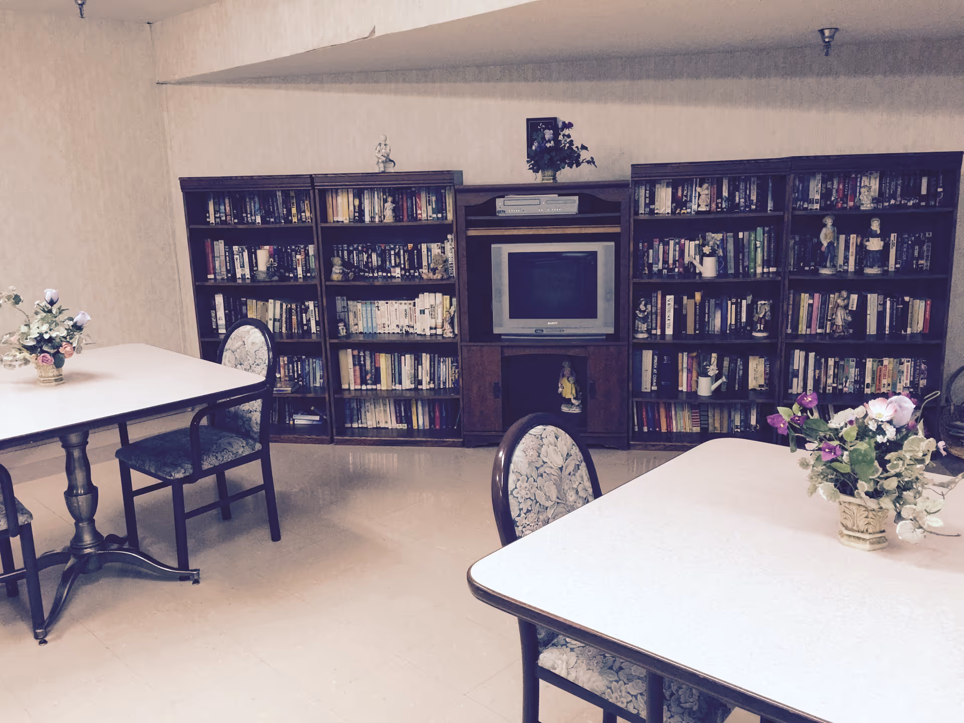 A senior living facility common area with bookshelves, a central TV, and tables with flower arrangements.