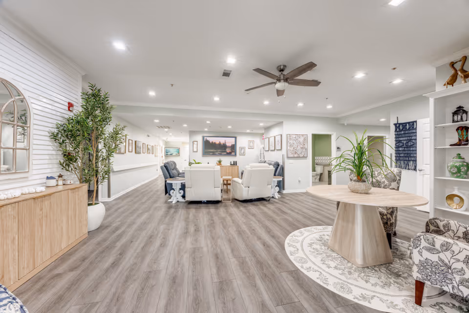Bright, spacious senior living lounge with sofas, a round table, and potted plants.