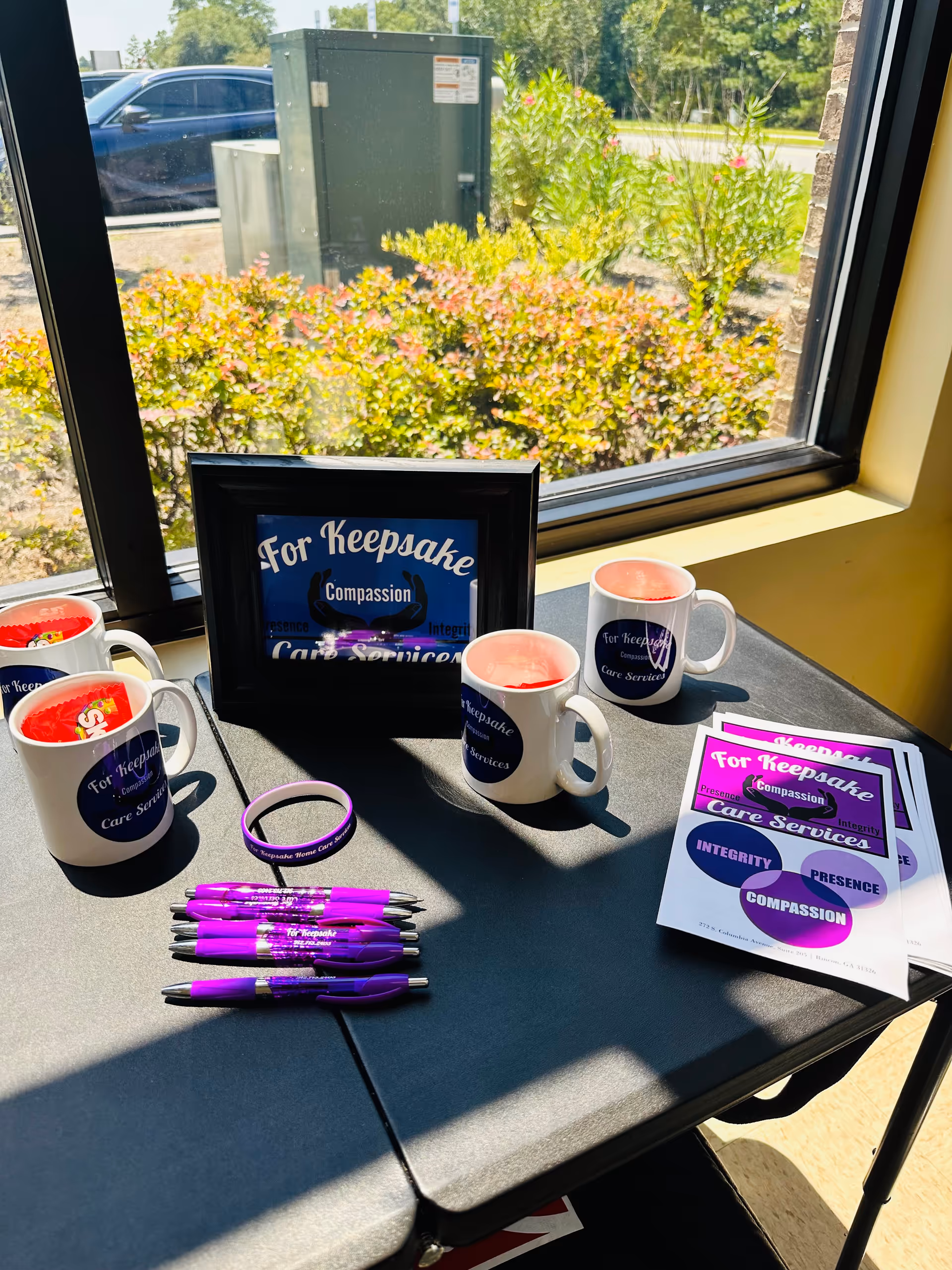 Promotional table with For Keepsake Care Services mugs, pens, and brochures by window.