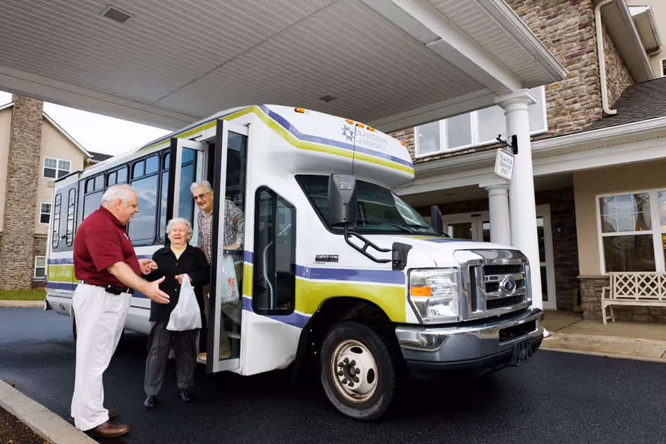 A shuttle bus parked at the covered entrance of a senior living facility while a staff member helps two elderly residents step off.
