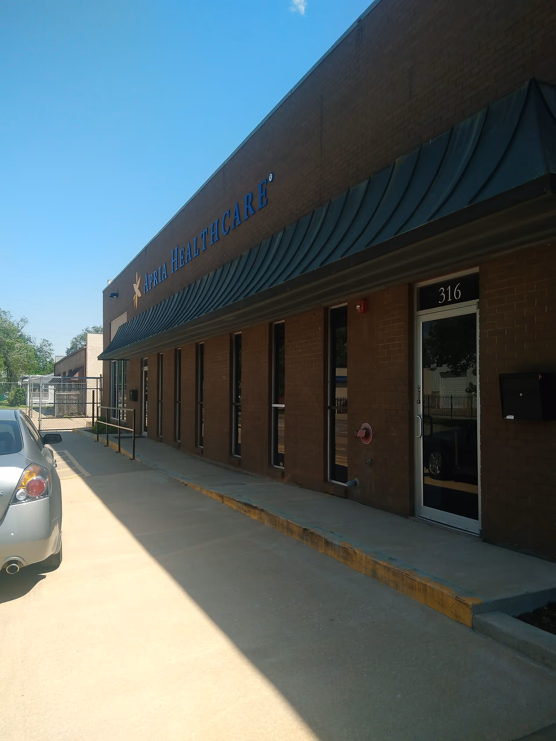 Exterior view of a brick Apria Healthcare facility with a glass entry door and parked car