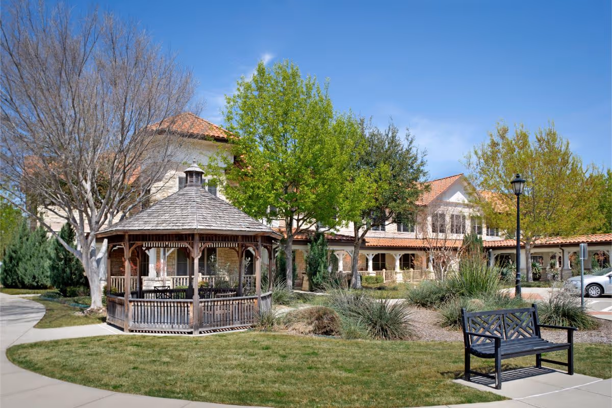 Front exterior of a senior living facility with a wooden gazebo, landscaped lawn, bench, and a two-story building with a covered porch.