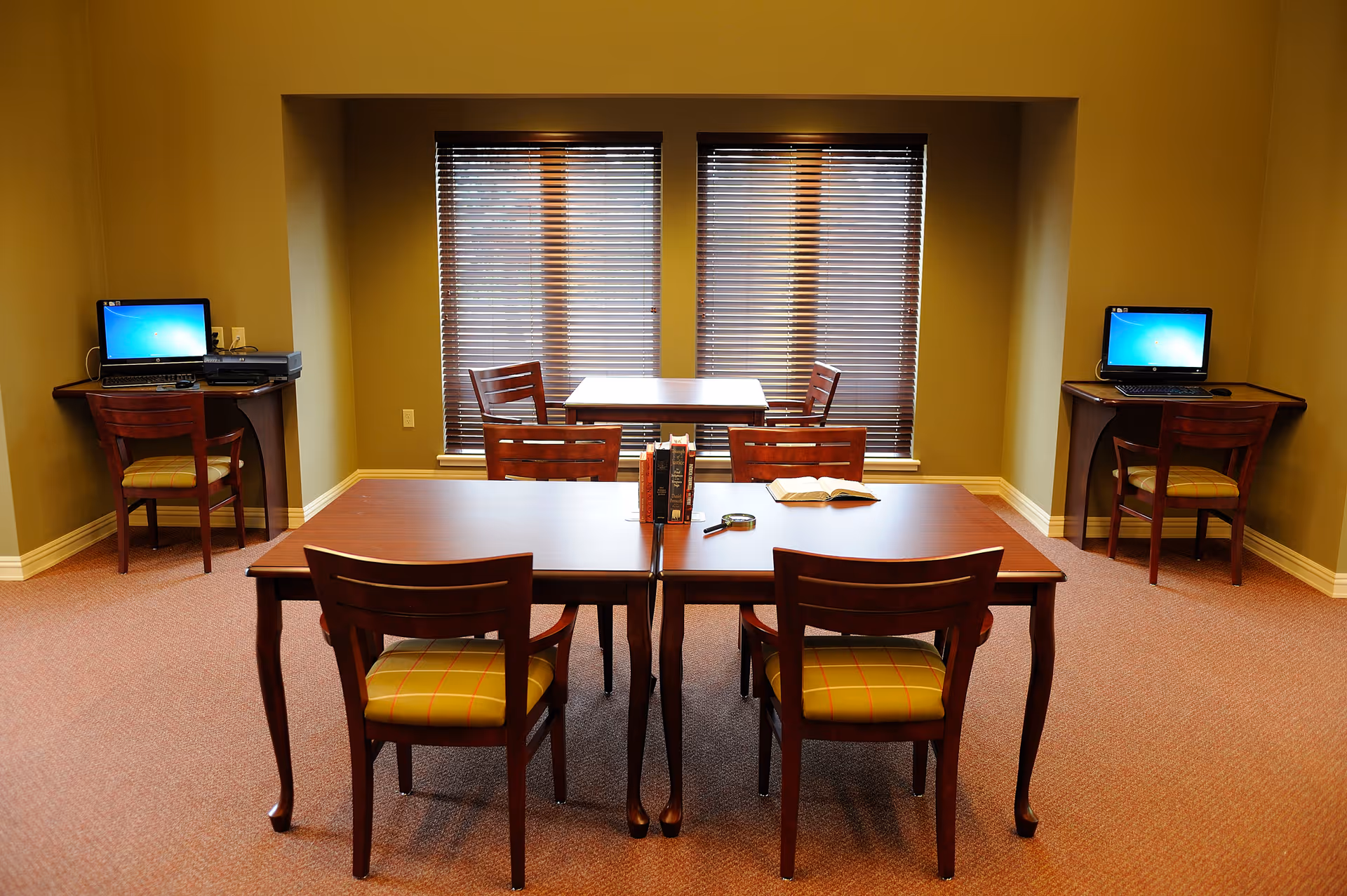 A communal interior room with a central wooden table, surrounding chairs, and laptops on side desks.