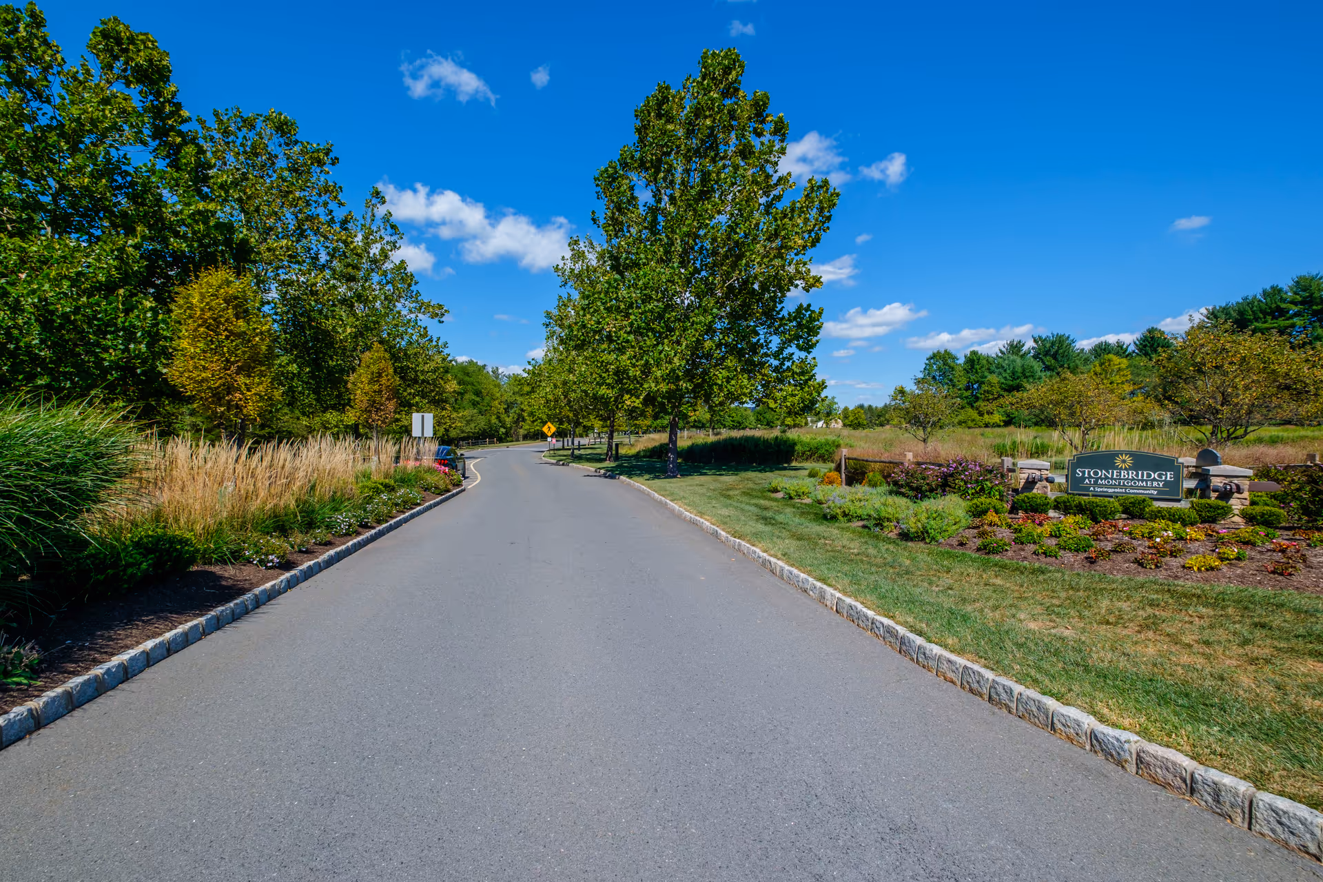 Tree-lined paved entrance road to Stonebridge at Montgomery with landscaped gardens and the facility sign on the right under a blue sky.
