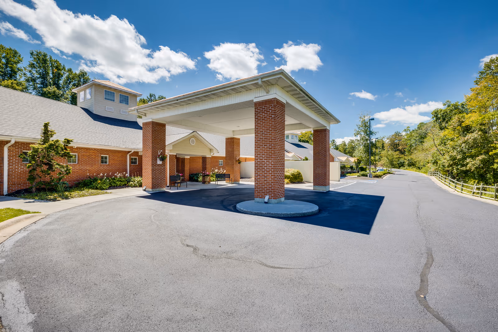 Entrance of a senior living facility with a covered drop-off area supported by brick columns, surrounded by a paved driveway and landscaped greenery under a blue sky with scattered clouds.
