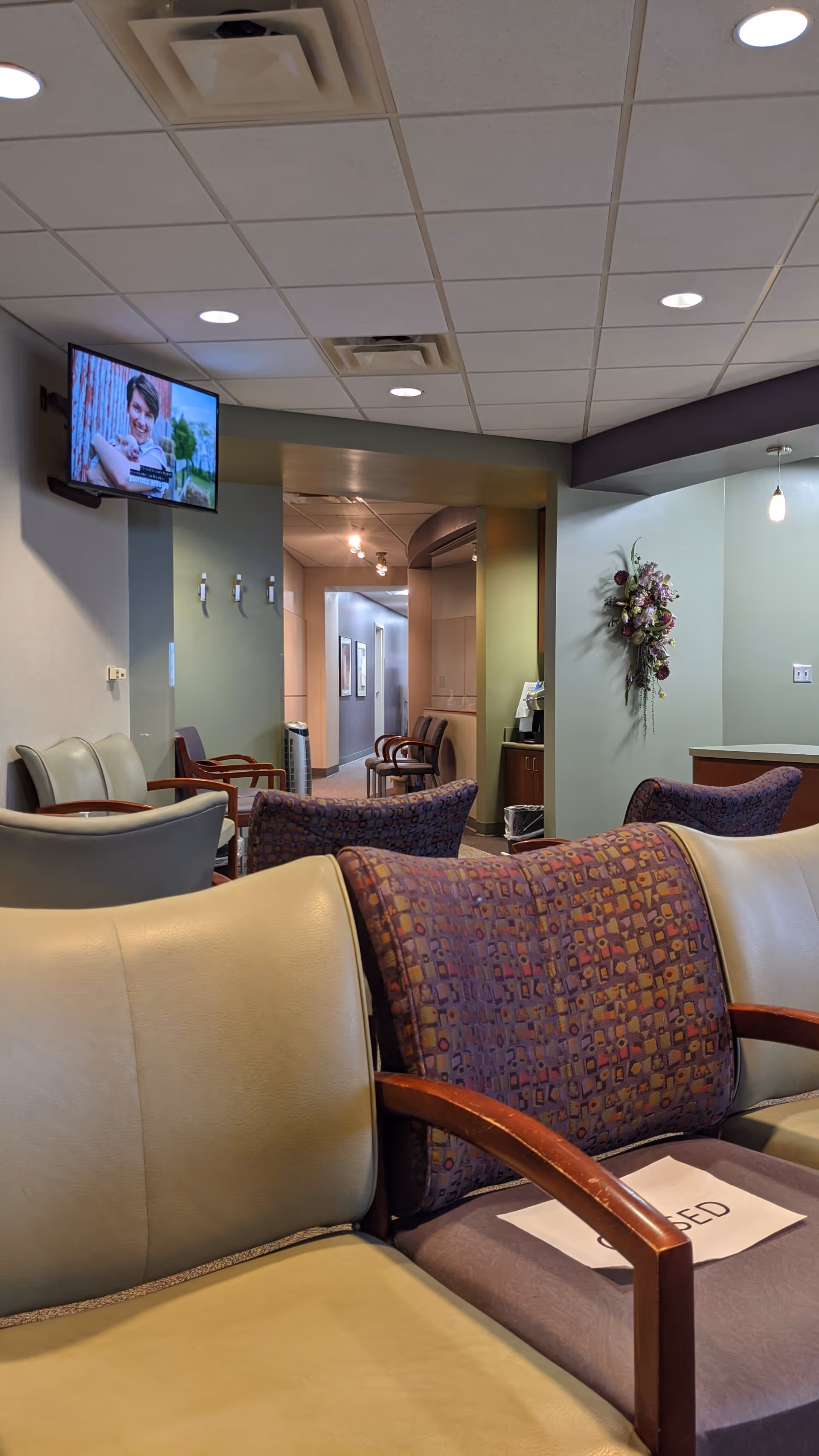 Senior living facility lounge with cushioned chairs, a wall-mounted TV, and a hallway beyond.