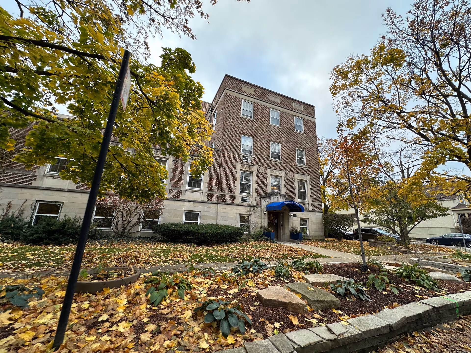 Exterior view of a multi-story brick building with a blue awning labeled Hartwell Place over the entrance. The building is surrounded by trees with autumn foliage and a landscaped area with fallen leaves and plants.