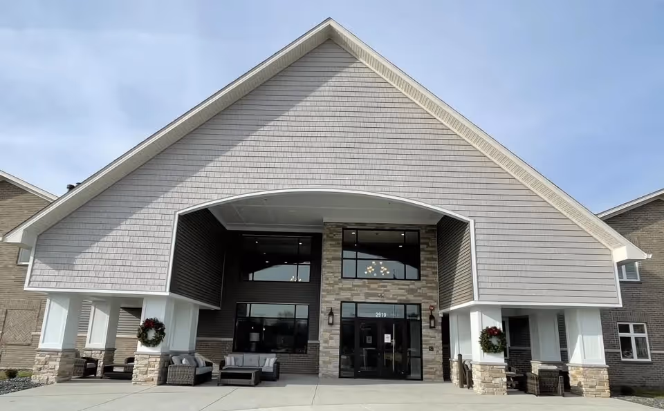 Front entrance of an assisted living building with a large gabled roof, stone columns, outdoor seating and holiday wreaths.