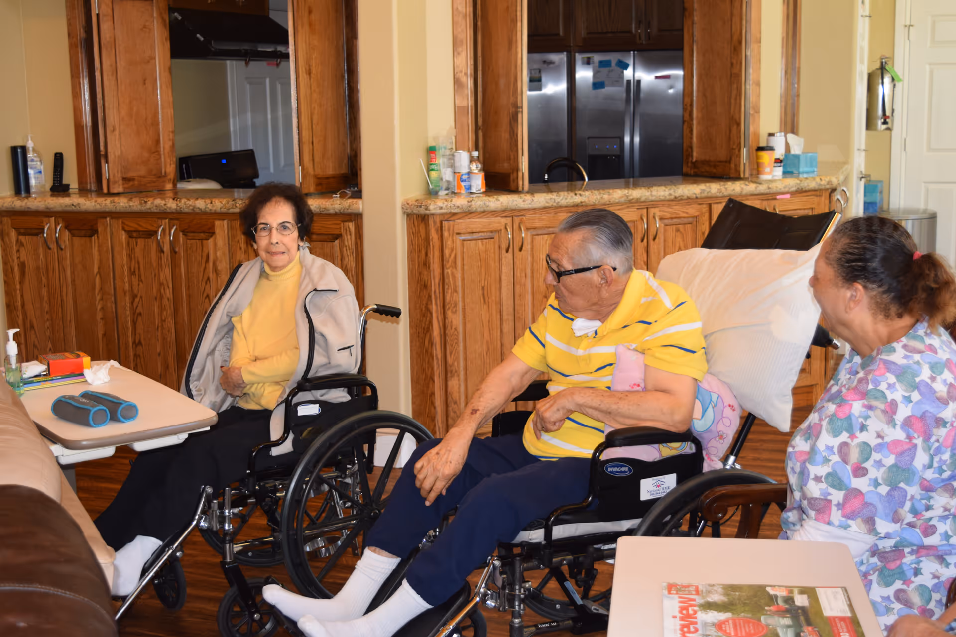Three elderly individuals in a living area of an assisted living facility. Two of them are seated in wheelchairs, one woman wearing a yellow sweater and a gray jacket, and a man in a yellow striped polo shirt with navy pants. A third person, possibly a caregiver, is sitting nearby wearing a colorful patterned shirt. The background shows wooden cabinets and a kitchen area with a refrigerator.