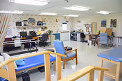 Well-lit multipurpose therapy/activity room with chairs, treatment tables, desks, and exercise/therapy equipment.