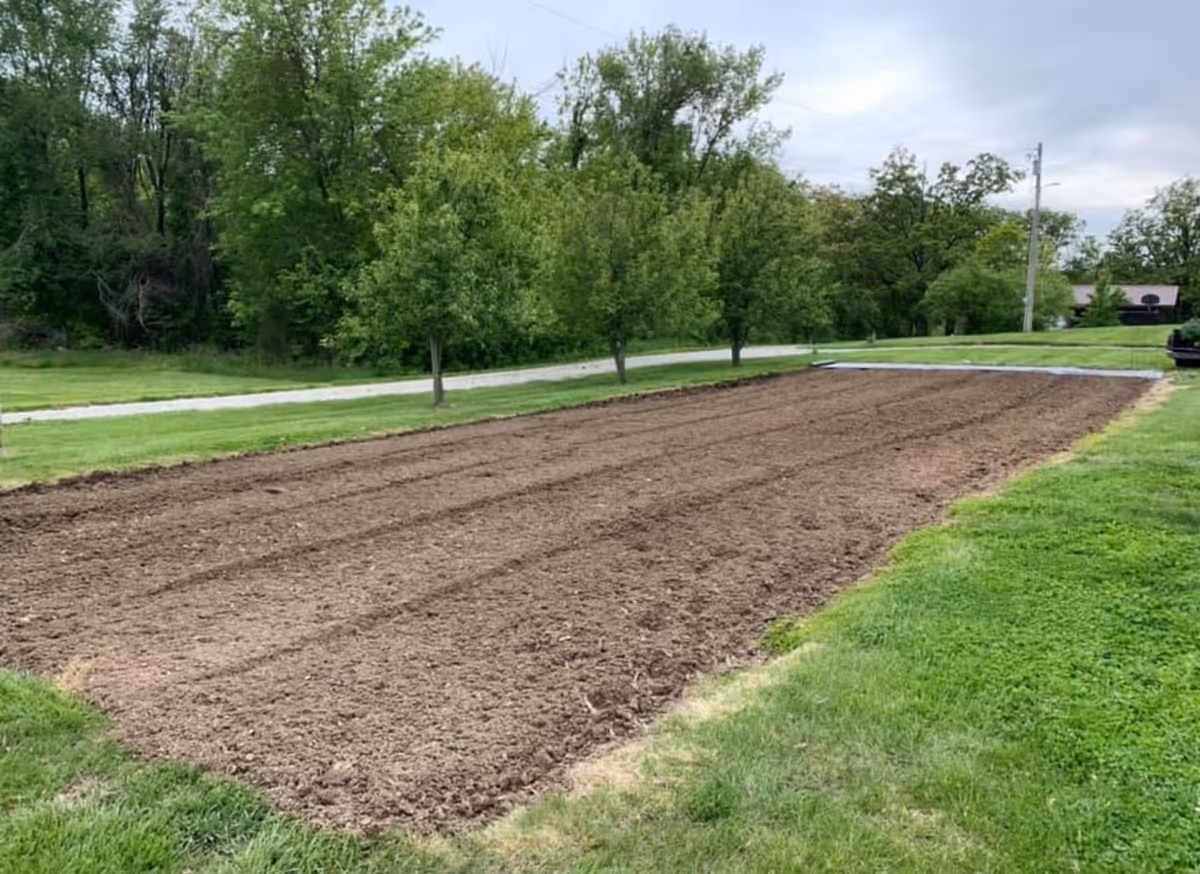 A freshly tilled rectangular garden plot surrounded by green grass with several trees and a road in the background under a cloudy sky.