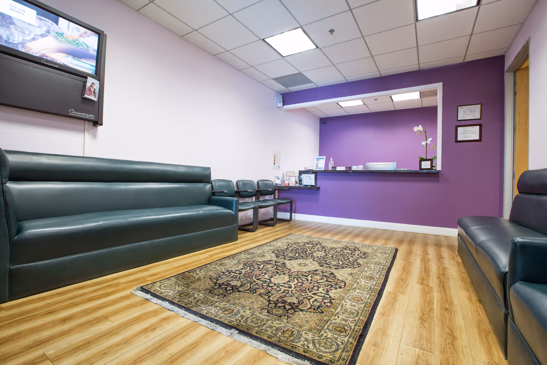 Modern waiting area with purple wall, leather sofas, chairs, a rug, and wall-mounted TV.