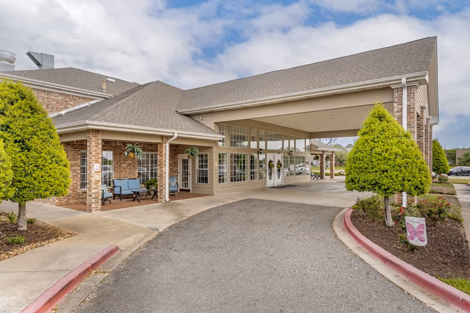 Entrance of a senior living facility with a covered driveway, brick exterior walls, large windows, and neatly trimmed trees and landscaping along the driveway.