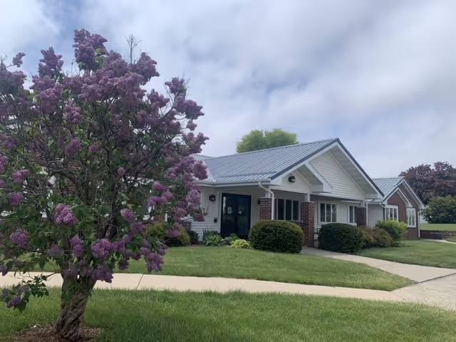 Single-story building with white siding and brick accents, featuring a metal roof. The building is surrounded by green grass, bushes, and a blooming purple lilac tree in the foreground. The sky is partly cloudy.