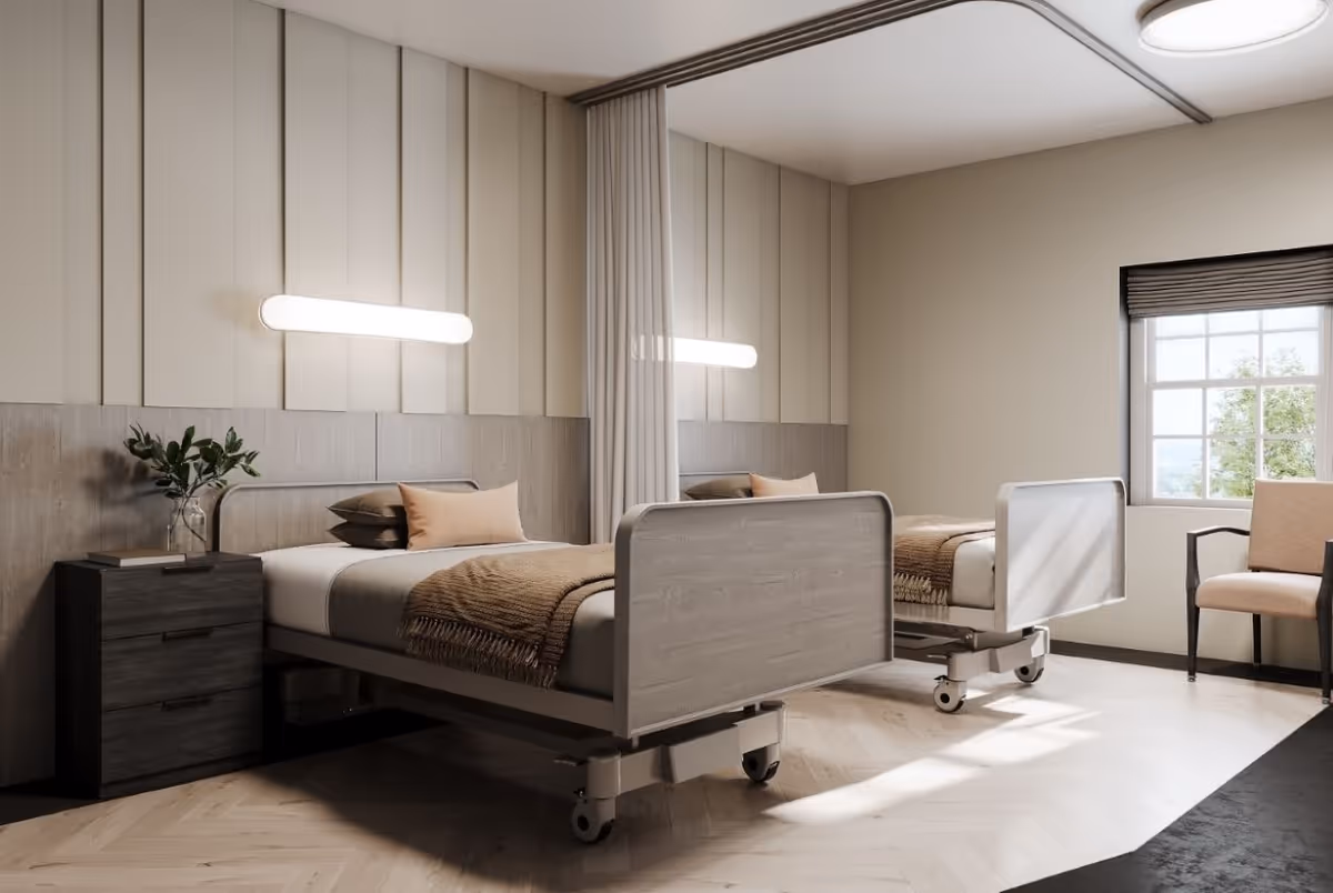 A bright and clean bedroom in a senior living facility featuring two hospital-style beds with wooden headboards and footboards, each made with beige and brown bedding. A small dark wooden nightstand with a glass vase holding green foliage is placed between the beds. A large window with a black blind allows natural light to fill the room, and a beige cushioned chair with black armrests sits near the window. The walls are painted in neutral tones, and a modern horizontal light fixture is mounted above the beds.