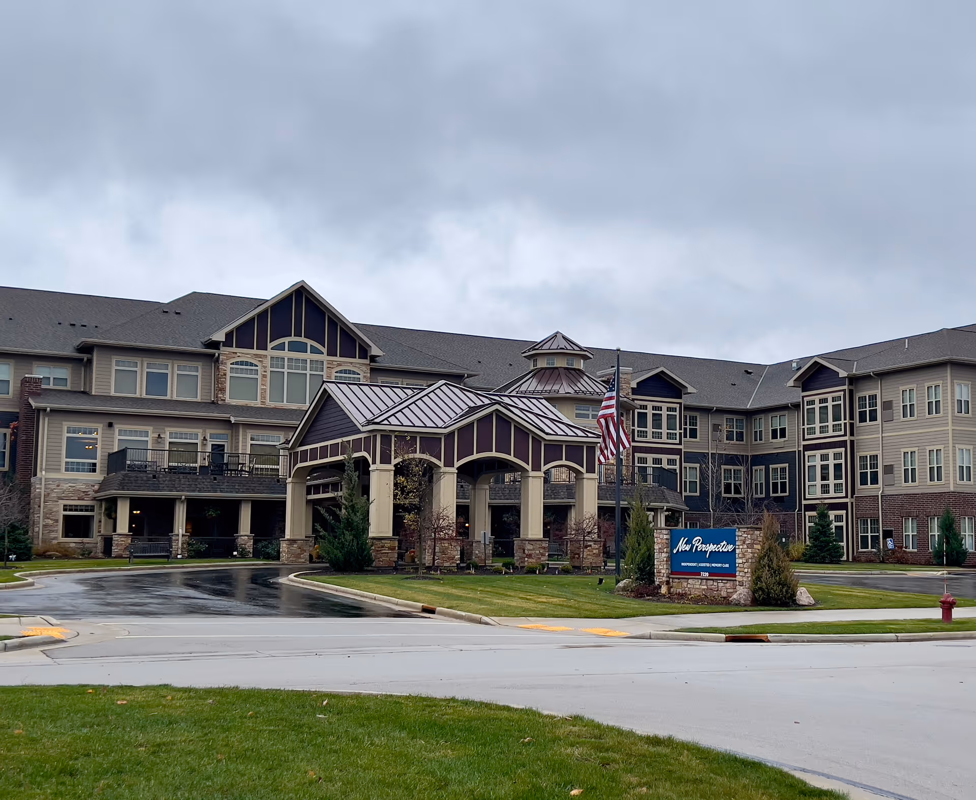Front exterior of a large multi-story senior living building with a covered porte-cochere, American flag, landscaped lawn, and a sign reading 'New Perspective'.
