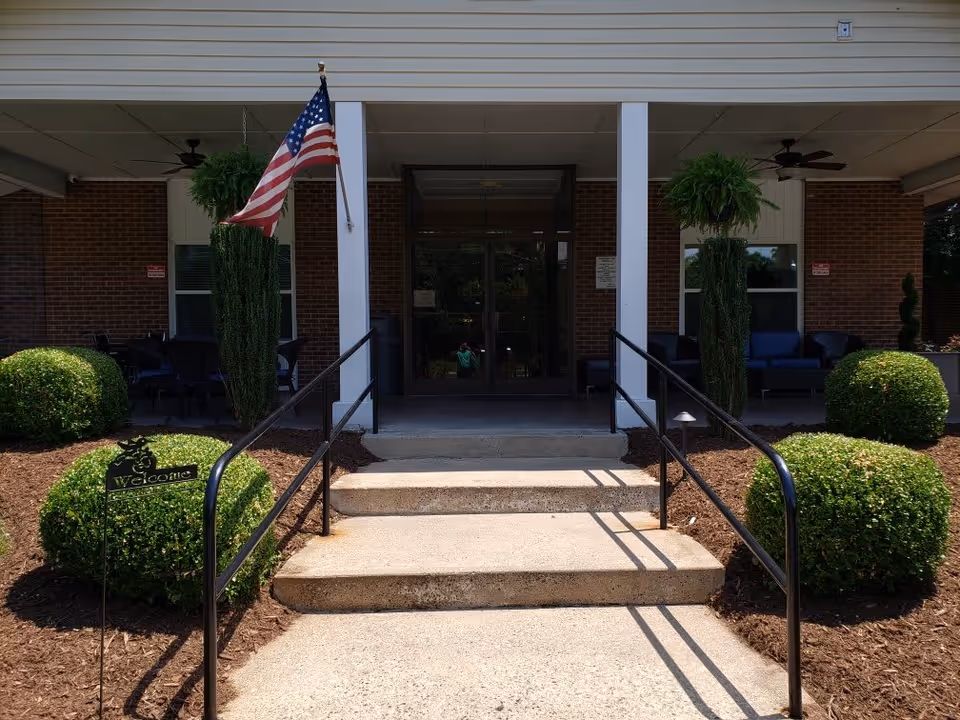 Entrance to a building with concrete steps and black handrails leading up to double glass doors. There are neatly trimmed bushes and two tall plants on either side of the steps, with an American flag mounted on the left side. The building has a covered porch area with ceiling fans and outdoor seating visible inside.