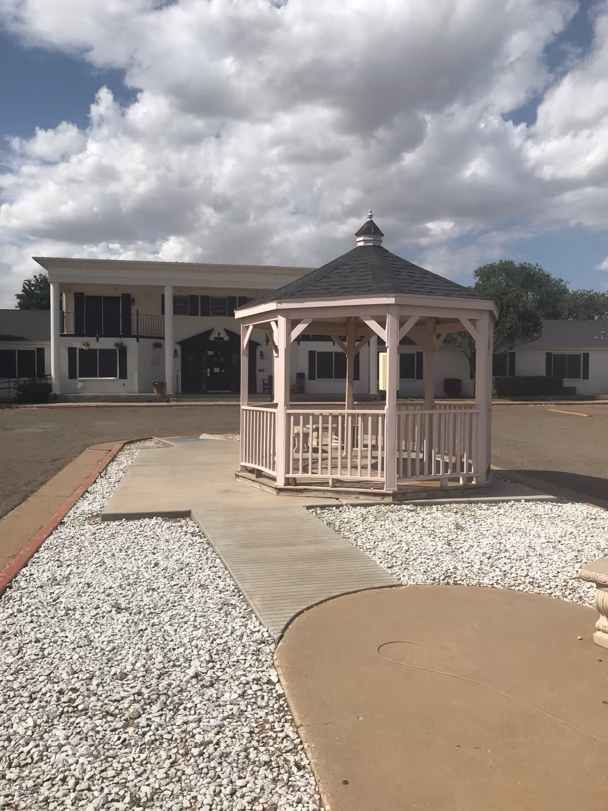 A light-pink wooden gazebo on a walkway in front of a two-story nursing facility building under a cloudy sky.