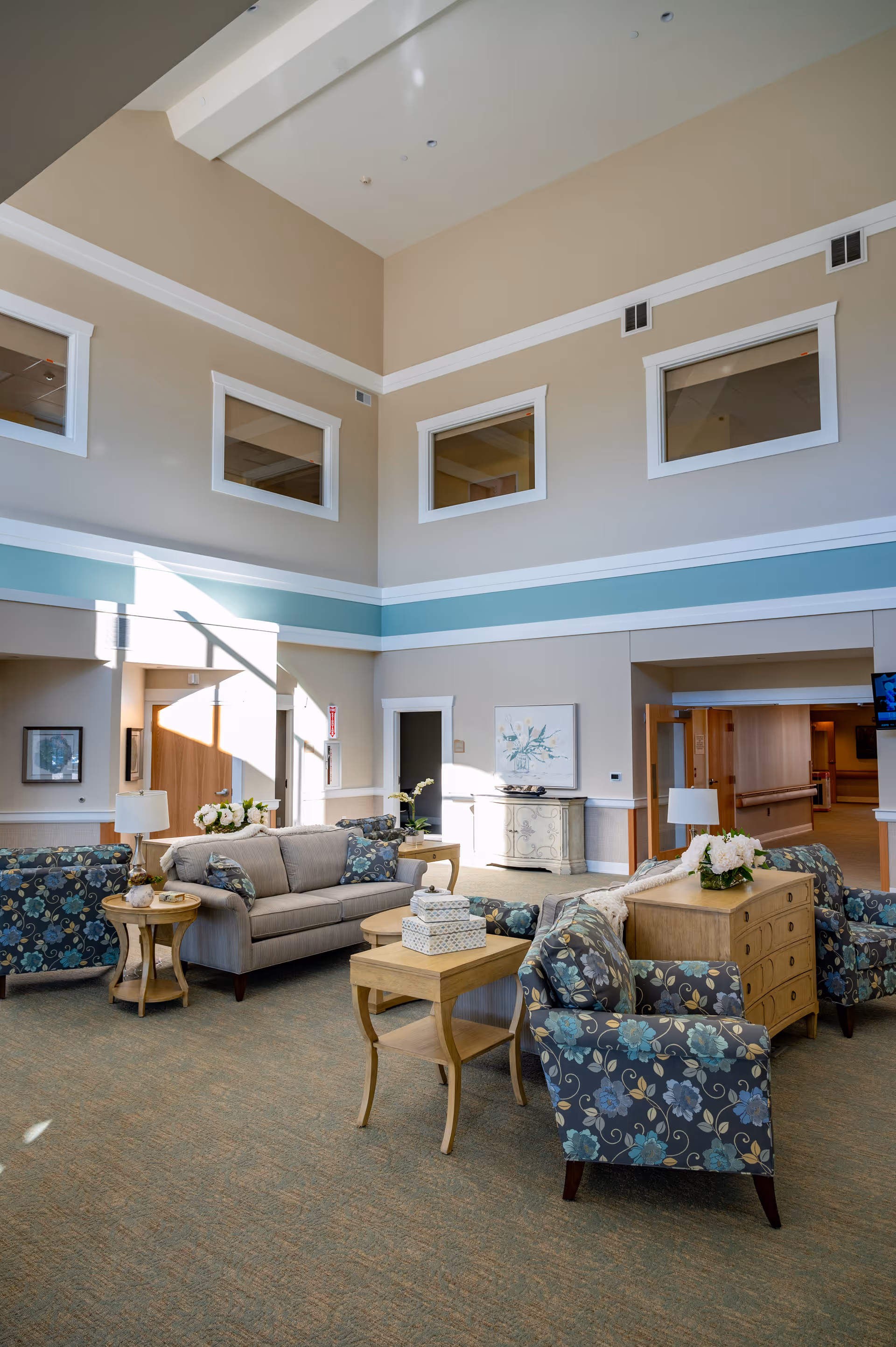 Bright, spacious lounge with sofas, floral armchairs, side tables and high ceilings in a senior living facility common area.