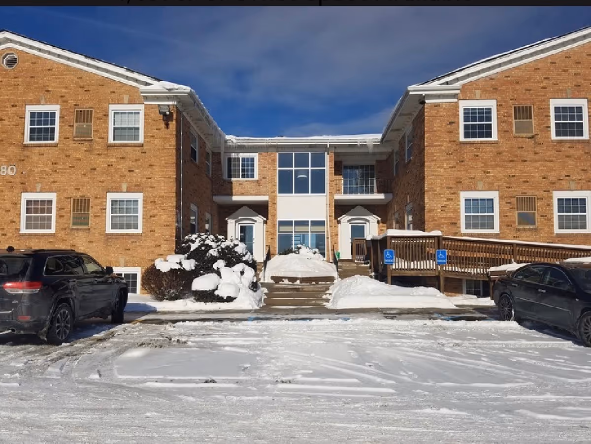 Brick senior living facility exterior with snow, stairs, and parked cars.