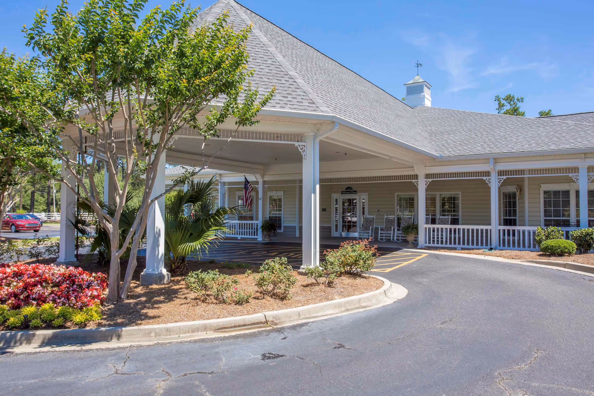 Exterior view of The Crossings of Georgetown facility entrance with a covered driveway, white columns, rocking chairs on the porch, landscaped flower beds, and a clear blue sky.