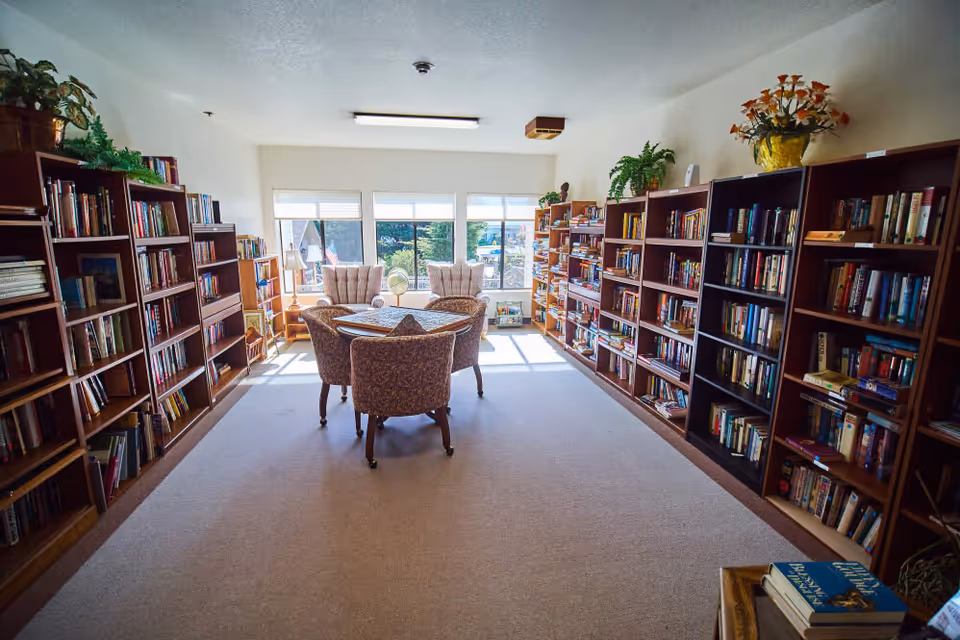 A bright and cozy library room with large windows letting in natural light. The room features multiple wooden bookshelves filled with books along both side walls. In the center, there is a round table covered with a cloth and surrounded by four upholstered chairs. Two armchairs are positioned near the windows, and there are plants and decorative items on top of the bookshelves.