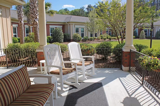 Covered patio with white rocking chairs and striped cushioned seating overlooking a landscaped courtyard.