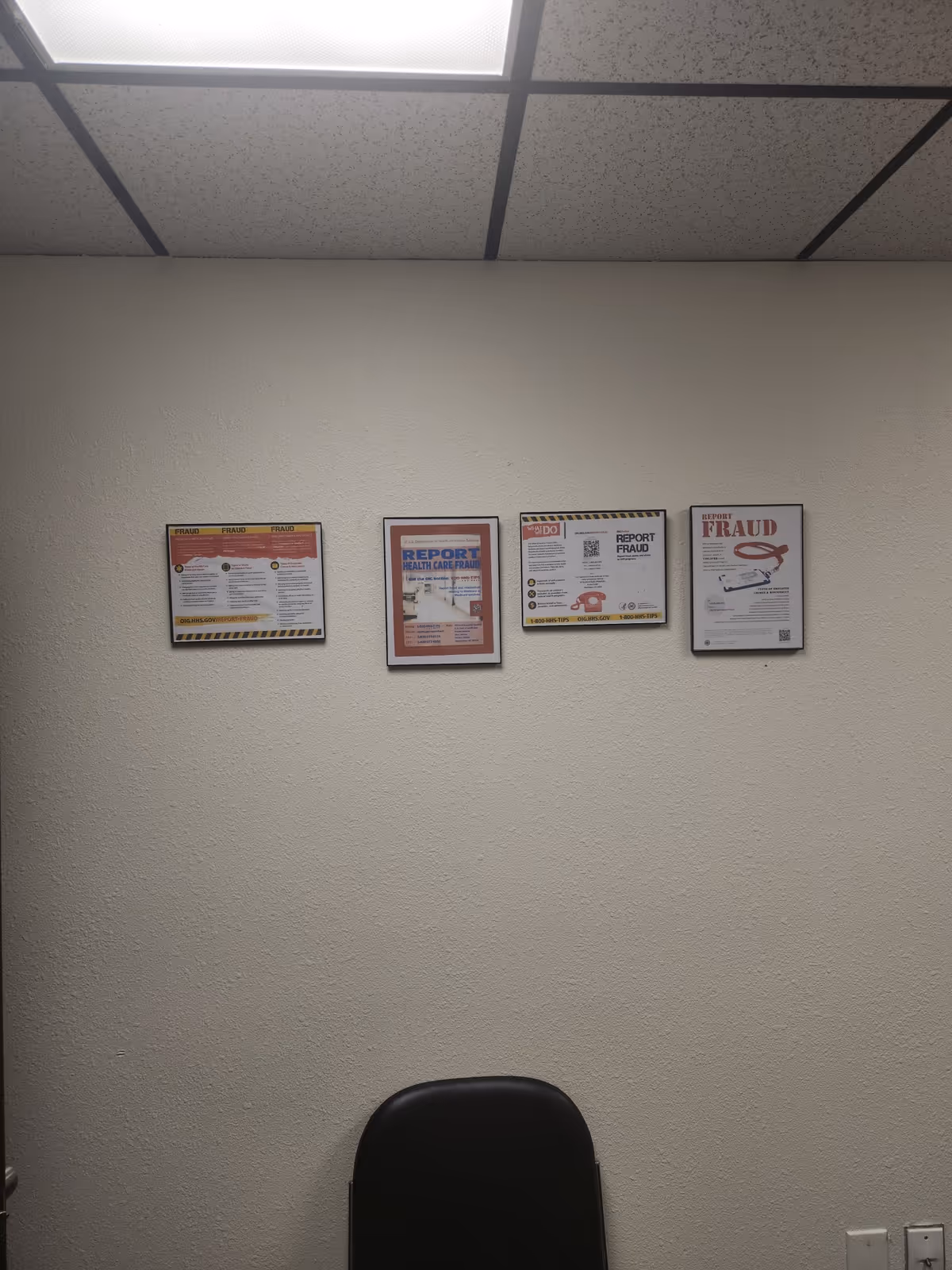 A plain hallway with beige walls, ceiling tiles, and framed posters above a black chair.