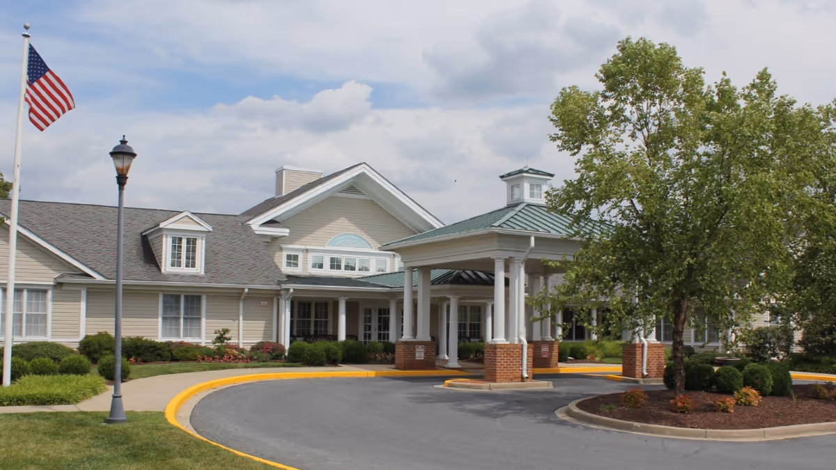 Front exterior view of Heart Fields Assisted Living At Frederick showing a single-story building with beige siding, white trim, a covered entrance supported by white columns with brick bases, a curved driveway, a flagpole with an American flag, a streetlamp, and landscaping including bushes and a tree.