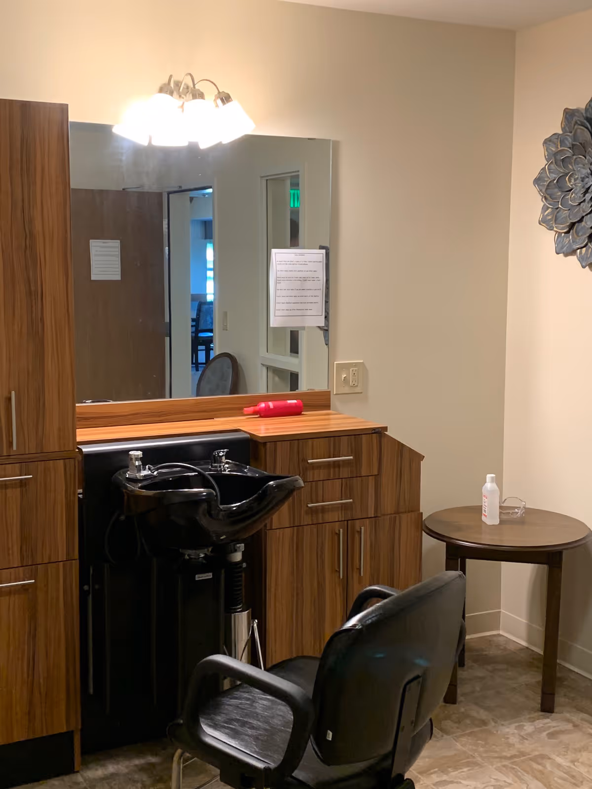 A salon area inside a senior living facility with a black salon chair in front of a black hair washing sink. The sink is set into a wooden cabinet with drawers and a countertop. Above the sink is a large mirror with a light fixture. To the right, there is a small round table with a bottle and a clear container on it. The walls are beige, and there is a decorative metal wall art piece on the right wall.