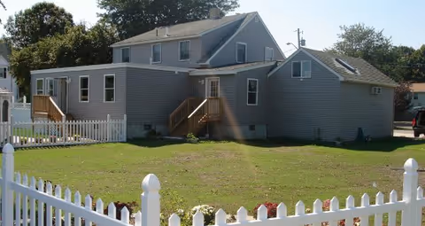 Exterior view of The Rubin Home with gray siding, stairs, and a white picket fence.