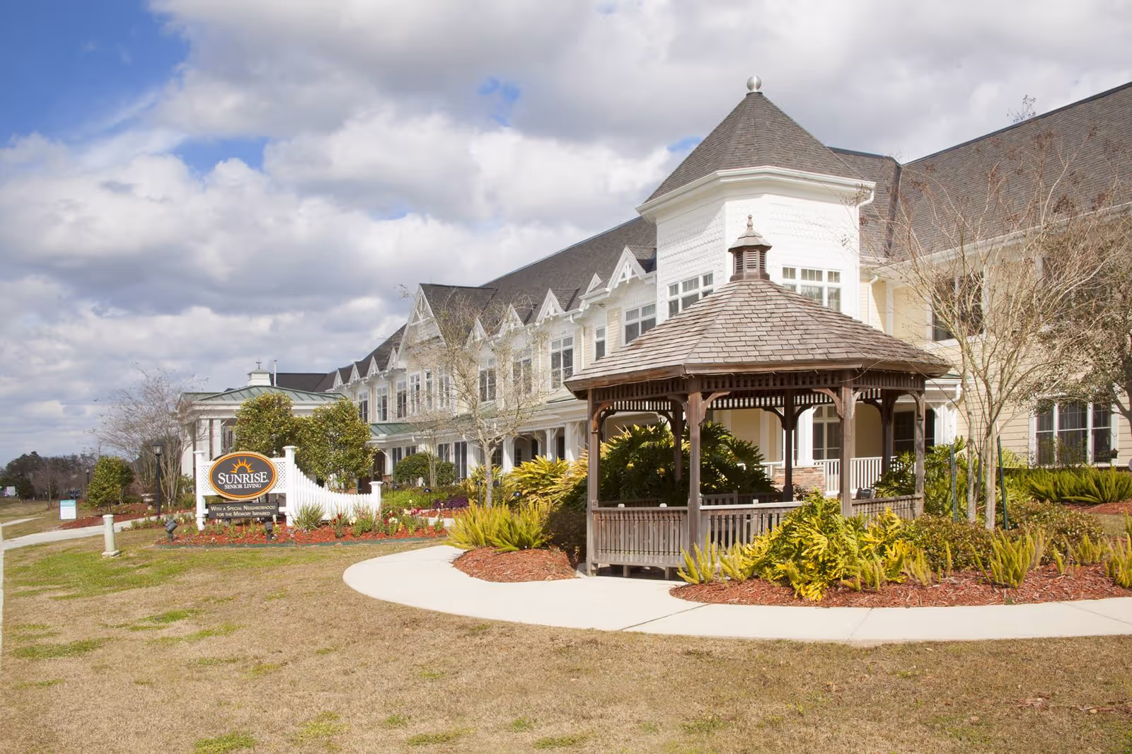 Exterior view of Sunrise at Siegen senior living facility featuring a large white multi-story building with a dark roof, a wooden gazebo surrounded by landscaped plants, and a sign with the facility's name near the entrance. The sky is partly cloudy.